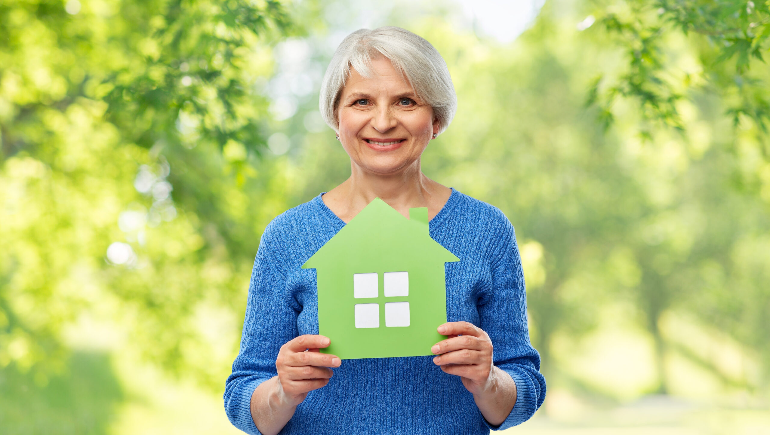 senior woman smiling holding a house sign with the trees behind her.